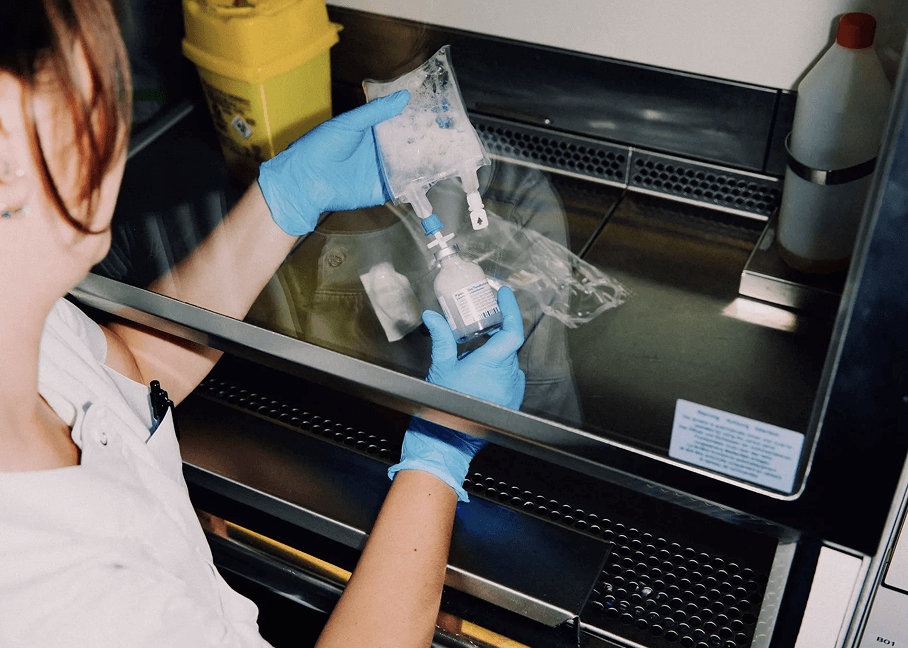 A person in gloves prepares an IV bag with medication in a sterile lab environment. Nearby are a yellow medical waste container and bottles.
