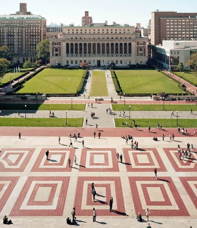 People walk across a plaza with geometric patterns leading to a grand building, surrounded by green lawns, with additional buildings in the background.