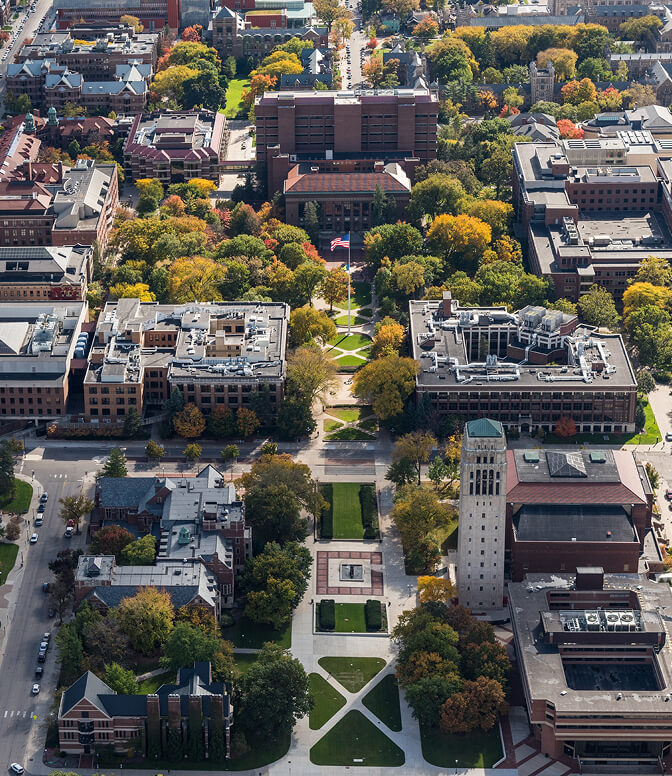 Aerial view of a university campus, featuring central grassy pathways converging at a flagpole, surrounded by academic buildings, trees with autumn foliage, and a prominent clock tower.