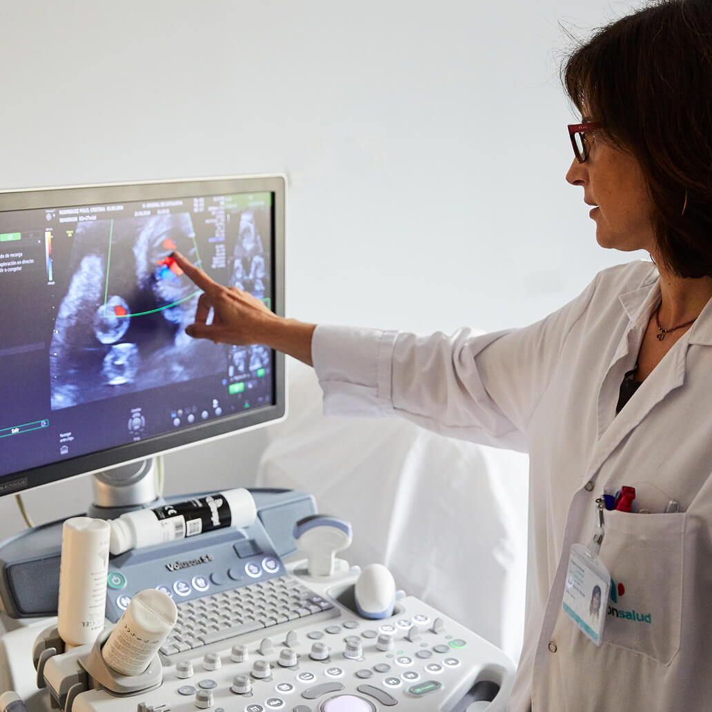 A healthcare professional points at an ultrasound image on a screen, highlighting details. The setting is a medical examination room with diagnostic equipment. Text on screen: unclear from the image provided.