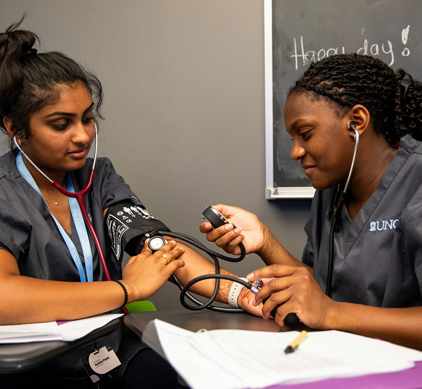 Two future medical students are practicing taking blood pressure in a classroom. One sits with a cuff on her arm, while the other uses a stethoscope and gauge. A chalkboard reads "Happy day!"
