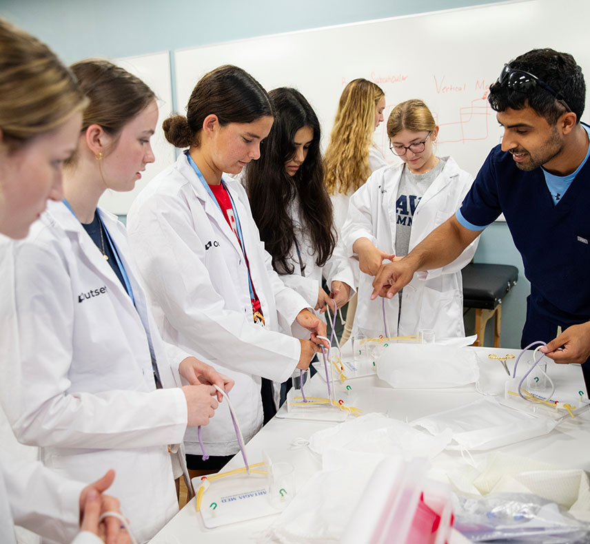 Students in white lab coats practice suturing techniques at a table, guided by an instructor in a dark shirt. The room has a light-blue wall and a whiteboard in the background.