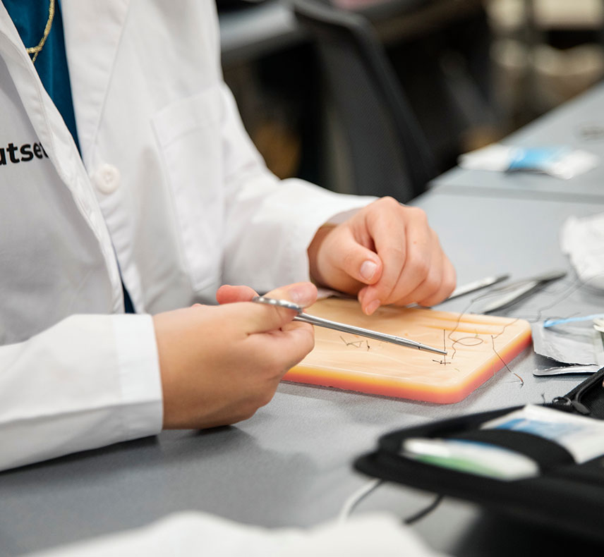 Hands hold a medical tool, practicing sutures on a synthetic skin pad. Student wears a white lab coat, likely in a clinical or educational setting with sterile instruments nearby.